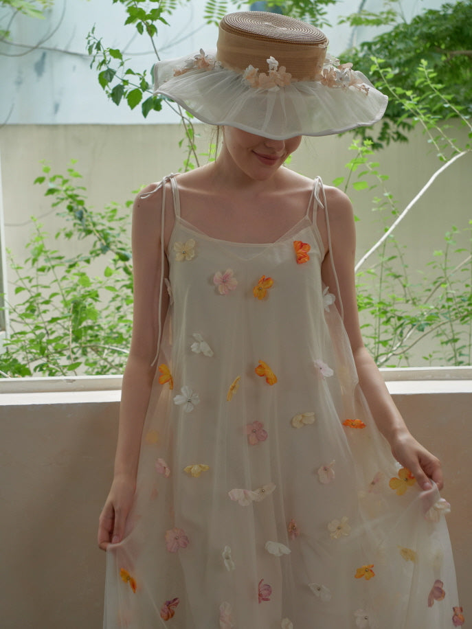 Wearing a dreamy floral mesh dress and a ruffled straw hat, the model smiles gently in a lush garden setting with fairytale spring vibes.
