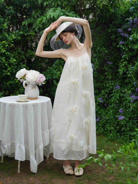 Model poses in a cream feather-embellished dress with thin straps and a wide-brim hat, evoking a whimsical fairycore mood in a garden setting.
