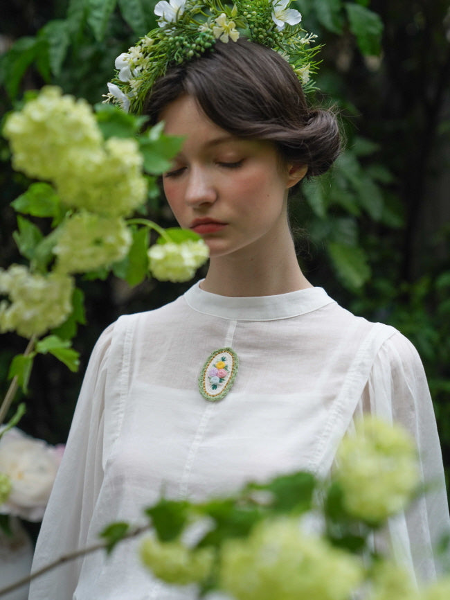 A young woman in a white vintage blouse with floral embroidery stands peacefully among soft green blooms, exuding vintage elegance.