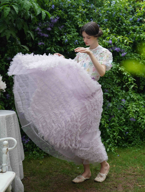The model lifts a voluminous lavender ruffle skirt while wearing a floral blouse, evoking a dreamy fairycore vibe in a garden scene.