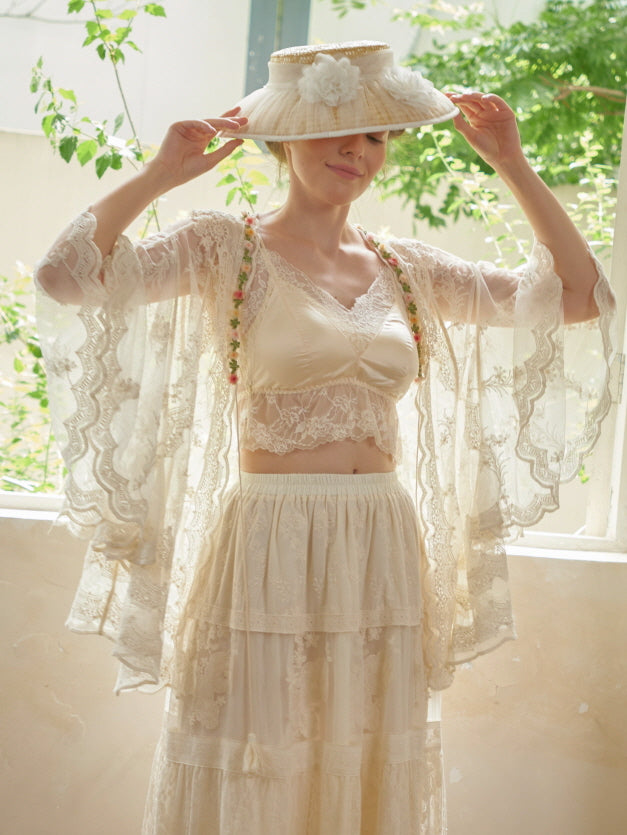 A woman stands in sunlight wearing a lacy fairycore robe over a satin bralette and embroidered skirt, styled with a floral hat.