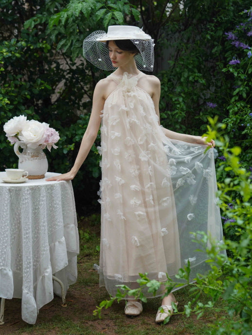 Wearing a romantic beige halter dress with sheer floral layers, the model stands by a lace table in a dreamy fairytale garden setting.