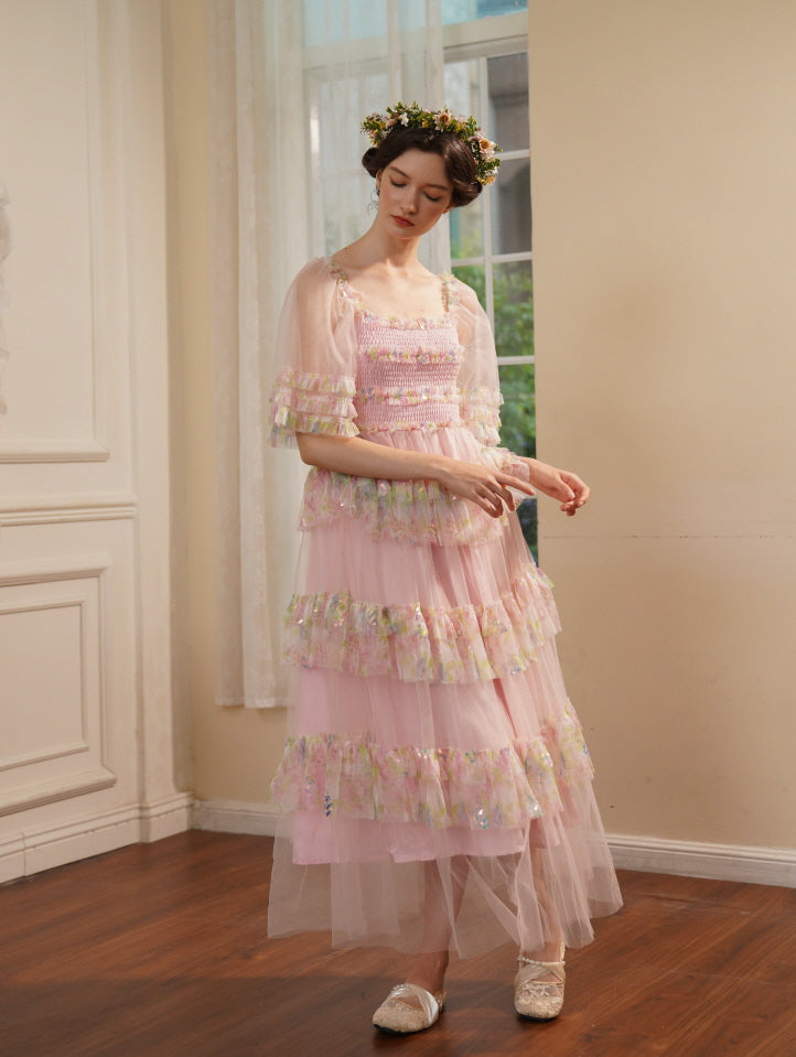 Wearing a layered pink dress with floral ruffles and sheer sleeves, a woman poses gracefully with a flower crown in a dreamy princesscore setting.