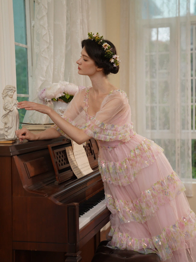 A woman in a layered pink fairycore dress leans over a piano, wearing a flower crown and gazing softly at a small statue in a romantic scene.