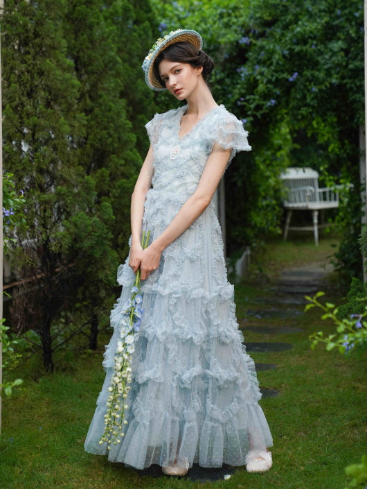Standing in a lush garden path, the model wears a tiered pastel blue gown with floral lace and puff sleeves, evoking a romantic princesscore dream.
