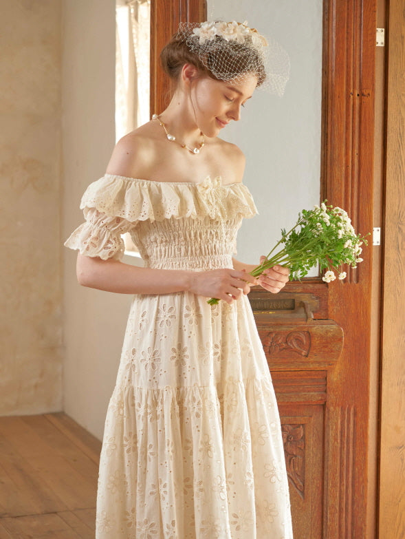 Standing beside a wooden cabinet, a woman in a princesscore lace dress gazes down at a bouquet with a soft and romantic expression.