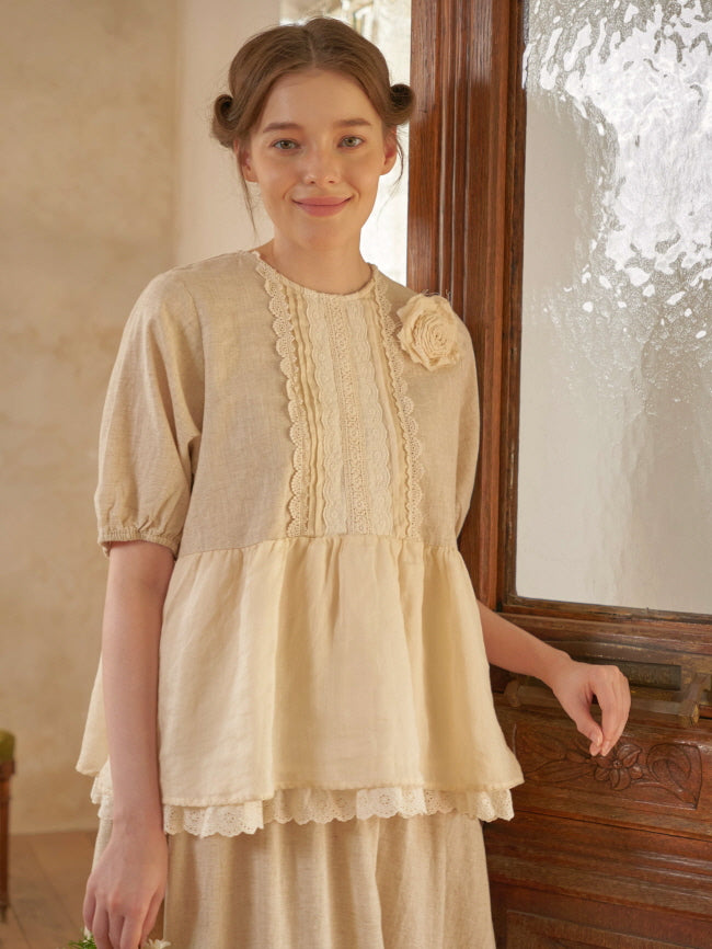 A smiling woman in a cream-toned cottagecore blouse with puff sleeves and a lace flower detail poses warmly beside an antique door.