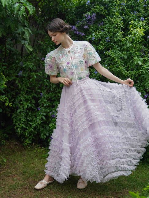 Wearing a pastel floral embroidered blouse and a tiered lavender ruffle skirt, the model poses gracefully in a lush garden fairycore scene.
