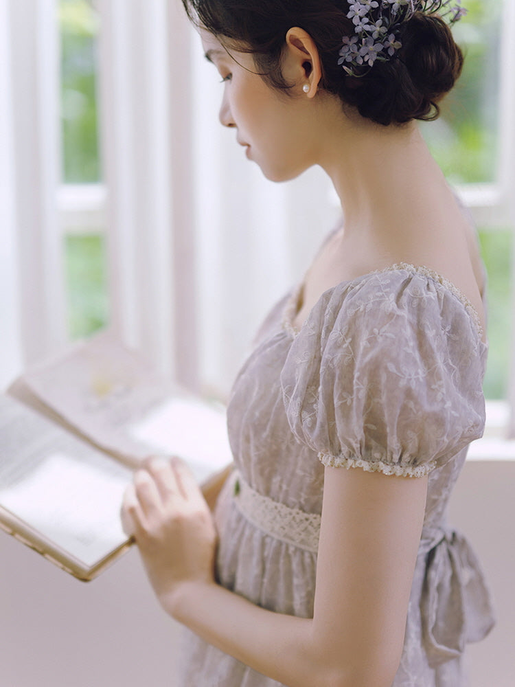 A woman reads a book by the window in a lavender puff sleeve dress with floral embroidery, radiating a peaceful princesscore mood.