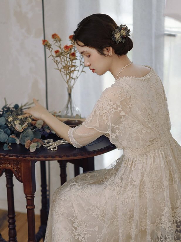 A woman in an elegant lace dress with floral embroidery sits in profile, arranging dried blooms on a vintage table in a romantic cottagecore scene.