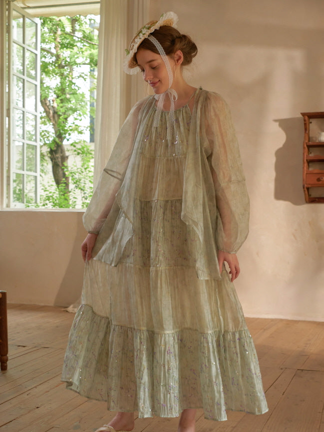 A woman in a sheer long-sleeve overlay and pastel tiered dress stands near a sunny window, radiating vintage fairycore charm.