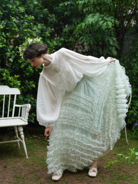 The model bends gracefully in a pastel ruffled skirt and white puff-sleeve blouse, surrounded by greenery in a dreamy fairycore garden.