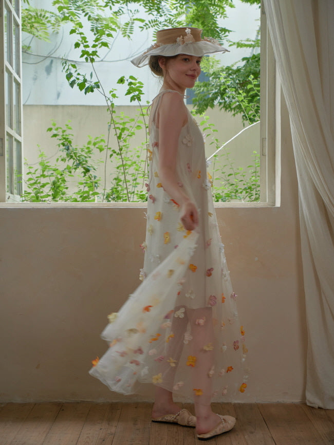 Turning with a gentle smile, the model showcases a fairytale-inspired flower dress and ruffled hat in a whimsical, nature-filled setting.