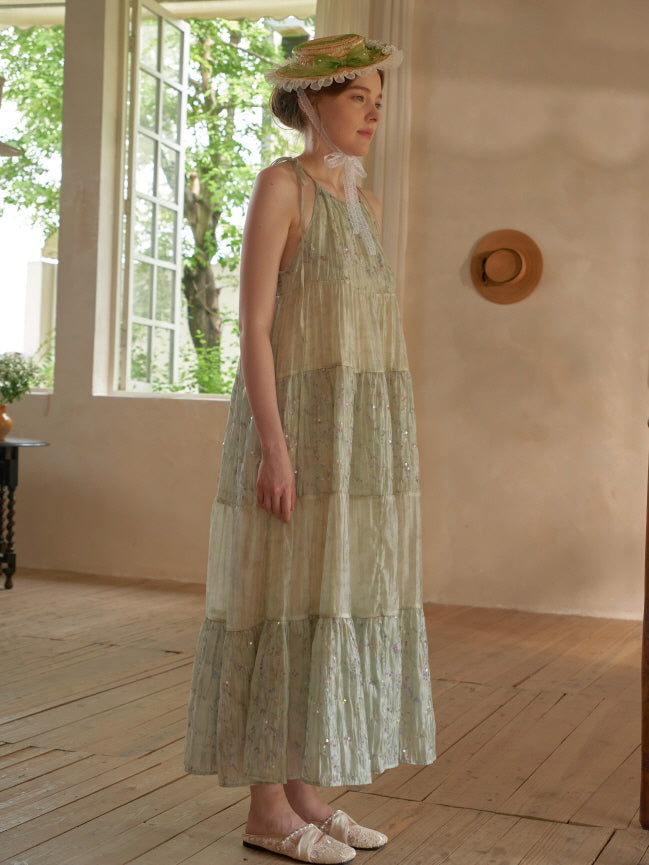 A woman gazes outside while wearing a pastel green halter dress with layered floral panels, styled with a bonnet in a vintage cottage room.