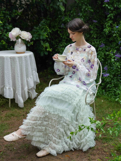 Seated in a white garden chair, the model sips tea in a sheer floral top and mint ruffle skirt, embodying soft princesscore elegance outdoors.
