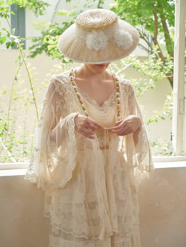 A close-up of a woman tying the ribbon of a sheer embroidered lace robe, styled with a floral strap and fairytale straw hat.