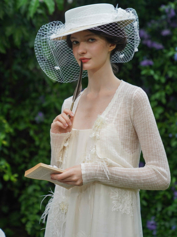 Model holds a feather and book while wearing a cream feather dress and sheer cardigan, creating a poetic cottagecore mood in a garden setting.
