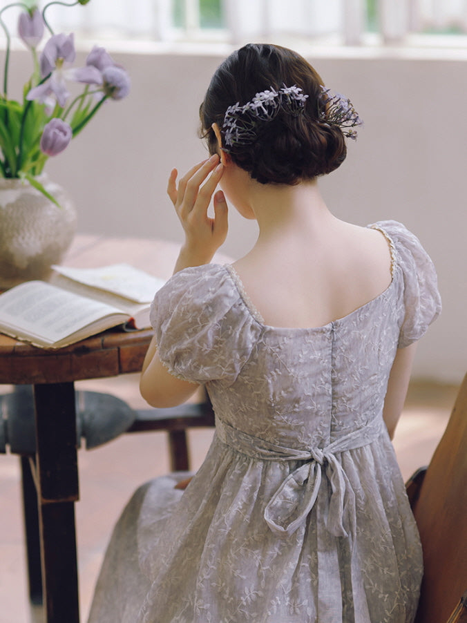 Seated at a vintage table, a woman in a lavender fairytale dress with a ribbon-tied back and floral hairpiece touches her ear softly.