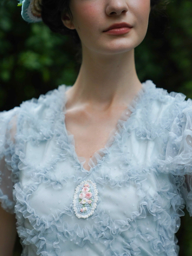Close-up of a pastel blue ruffle fairytale dress with a delicate embroidered floral brooch, highlighting the romantic vintage-inspired texture and details.
