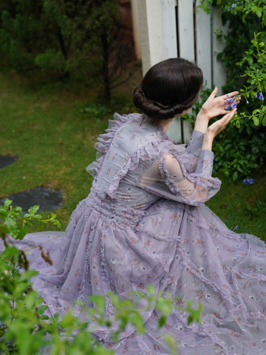 Seated on the grass in a vintage-inspired lavender dress, the model holds delicate flowers in a peaceful princesscore garden setting.