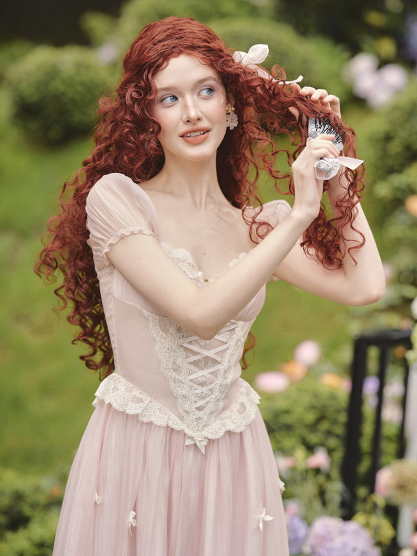 A red-haired woman brushes her curls in a pink corset dress with lace trim, capturing a whimsical fairytale moment in a cottagecore garden.
