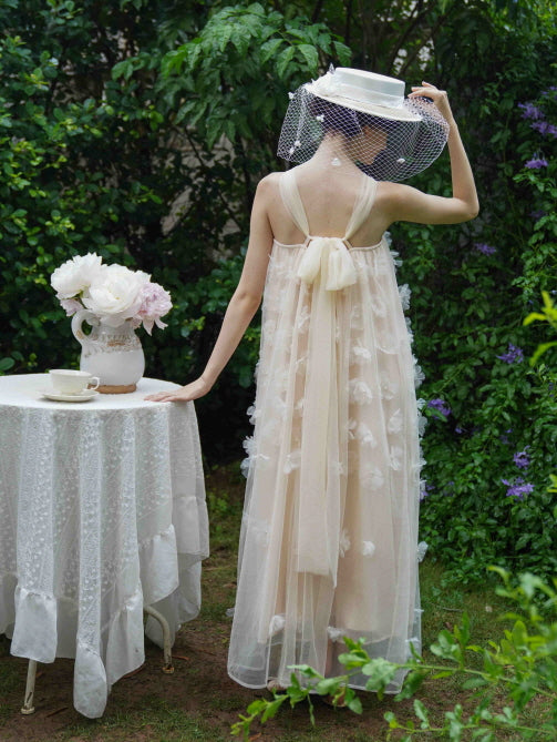 Back view of a model in a sheer beige halter dress with a long ribbon bow, standing by a floral table in a vintage-inspired garden setting.