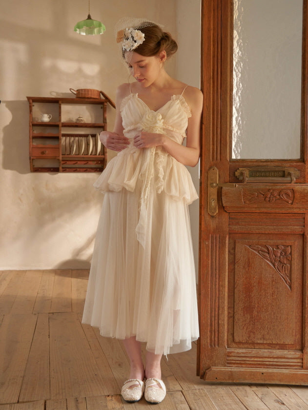 A woman gently looks down while wearing a cream tulle dress with ruffle details and floral waist accent, styled in a dreamy cottagecore room.