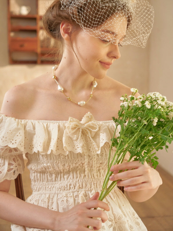 A woman with a floral veil and pearl necklace smiles softly while holding chamomile flowers, dressed in a romantic off-shoulder eyelet dress.