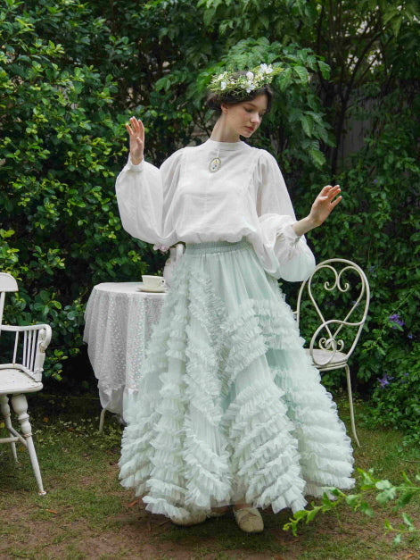 Wearing a flowing mint ruffle skirt and vintage white blouse, the model poses elegantly in a lush garden for a princesscore-inspired look.