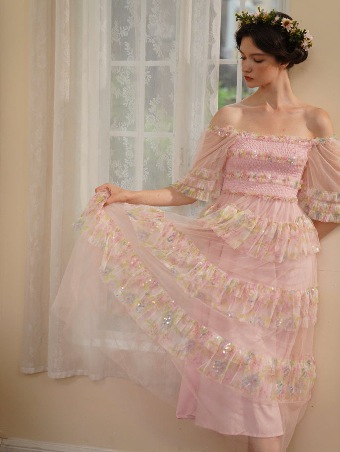 Wearing a pastel pink layered dress with sequined floral ruffles, the model gently holds out the sheer skirt in a graceful princesscore pose.