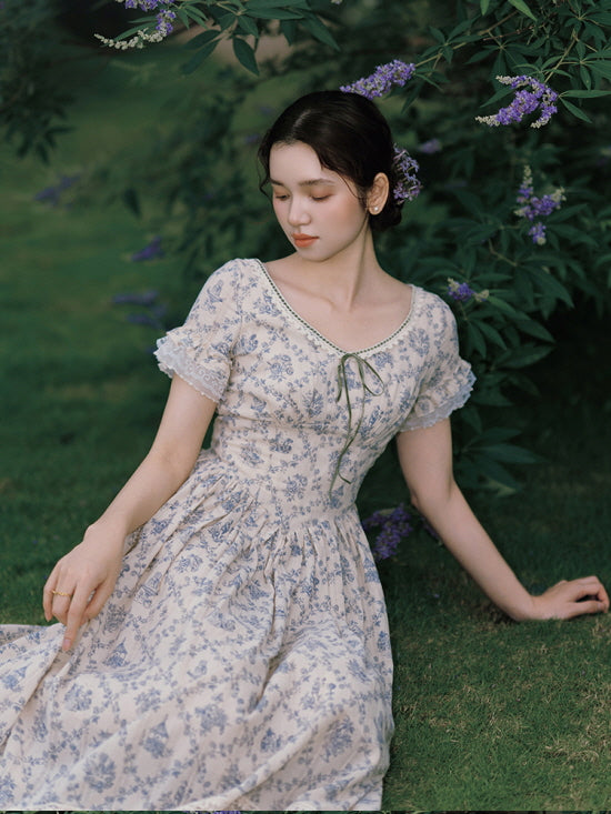 The model reclines gracefully on the grass, wearing a soft, cottagecore-style dress adorned with blue floral patterns and lace trim, evoking a dreamy, fairytale ambiance.