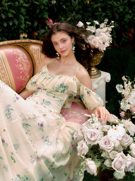 Leaning back among blush roses, the model lounges in a ruffled floral dress with dreamy off-shoulder sleeves, perfect for a fairycore summer aesthetic.