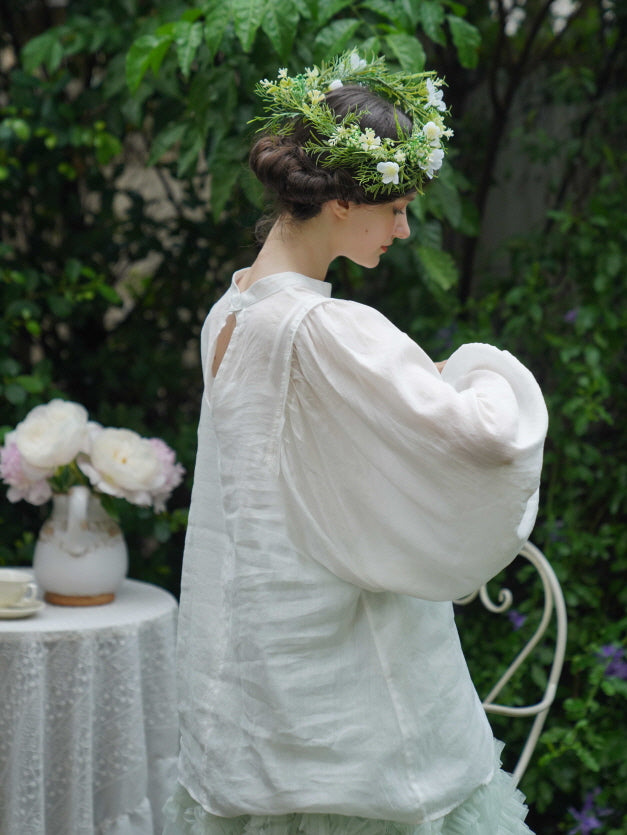 A woman in a white puff-sleeve blouse and pastel ruffle skirt stands in profile, her hair crowned with wildflowers in a romantic vintage garden scene.