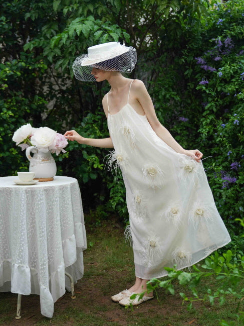 Wearing a cream dress with feather florals and a wide-brim hat, the model leans toward a tea table in a whimsical fairycore-inspired pose.