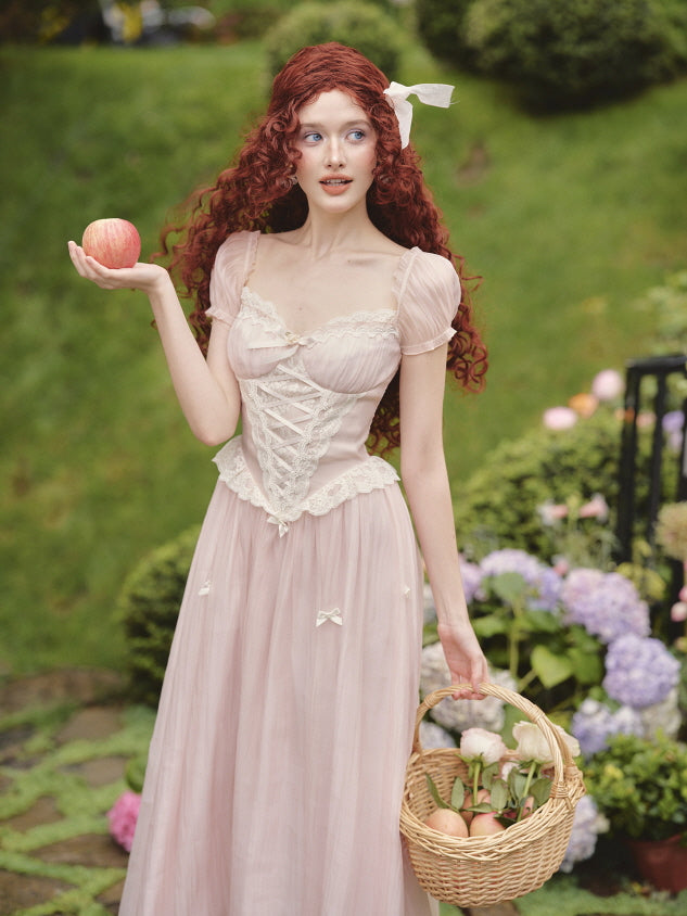 Model holds an apple and a basket in a blush princesscore dress with puff sleeves and lace-up bodice, styled in a dreamy floral setting.