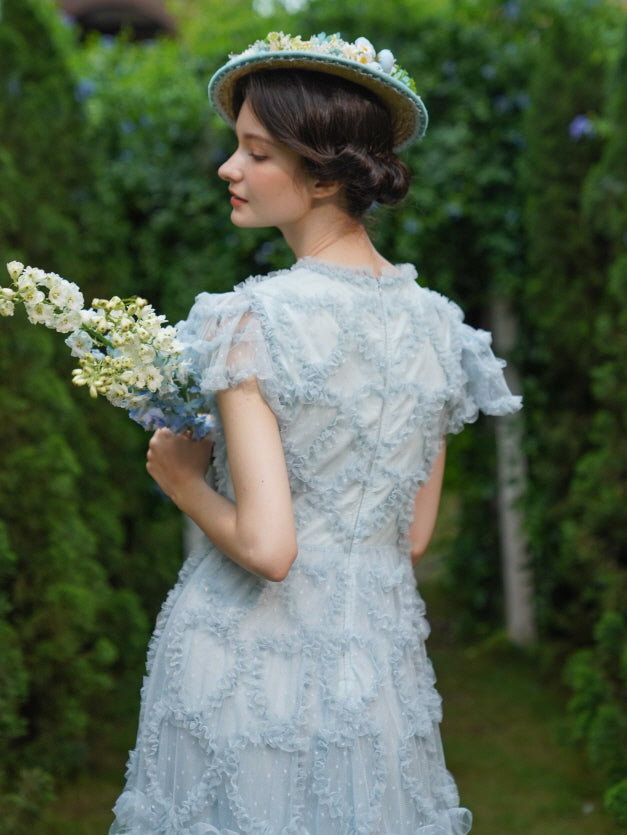Model holding soft wildflowers with her back turned, showcasing the airy ruffled back of a fairytale dress in a dreamy garden setting.