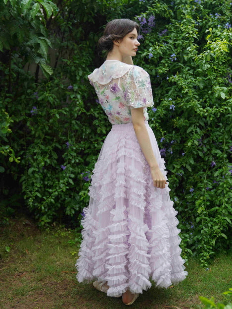 Back view of a woman in a pastel floral blouse with lace collar and a lavender layered skirt, styled in a fairycore garden setting.