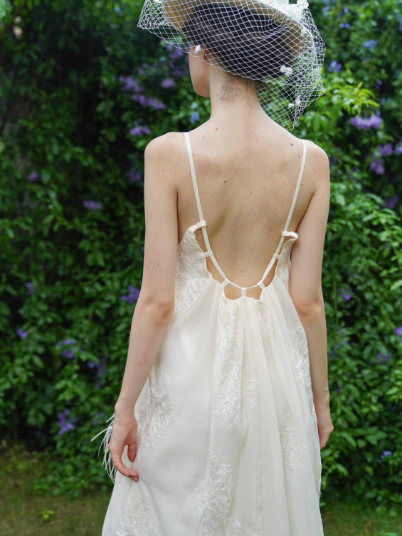 Back view of a cream feather dress with a low-cut strappy design and embroidered detail, styled with a romantic netted hat in a garden.