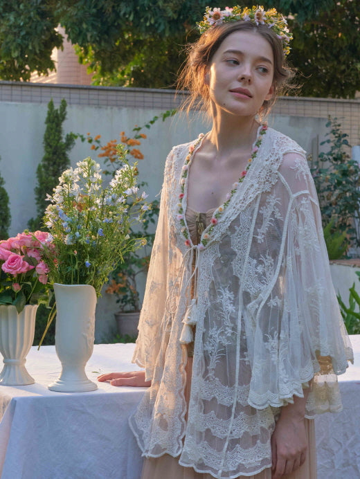 A woman in a sheer lace cape over a corset dress leans beside a floral table, styled with a flower crown for a romantic fairycore look.