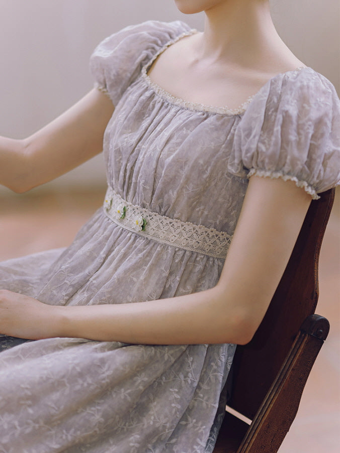 Close-up of a romantic lavender dress with puff sleeves and daisy-accented lace waist, worn by a woman seated in vintage fairytale elegance.