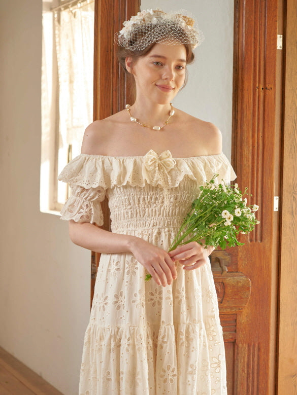 Wearing a lace ruffle dress and holding a small bouquet, the woman stands by a vintage door in a sunlit room with princesscore elegance.