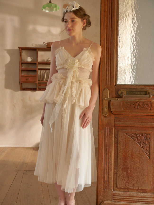 Standing gracefully by a wooden door, she models a soft, layered cream dress with floral lace, reflecting a dreamy fairytale aesthetic.