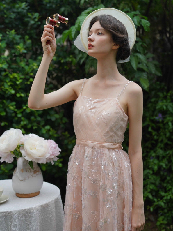 Wearing a soft pink embellished dress with pearl straps, the model lifts opera glasses in a garden, capturing vintage elegance and princesscore charm.