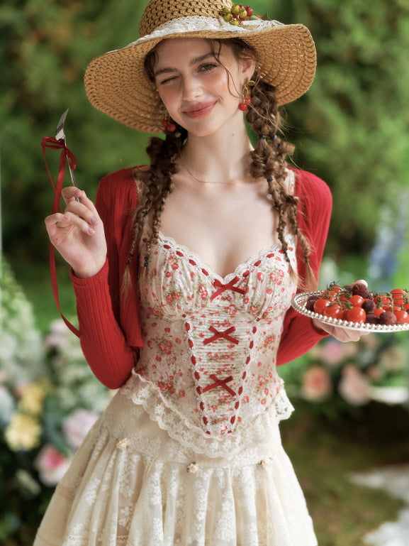 Model smiles in a floral corset top with red ribbon detail, styled with a straw hat and cardigan for a sweet coquette cottagecore look.