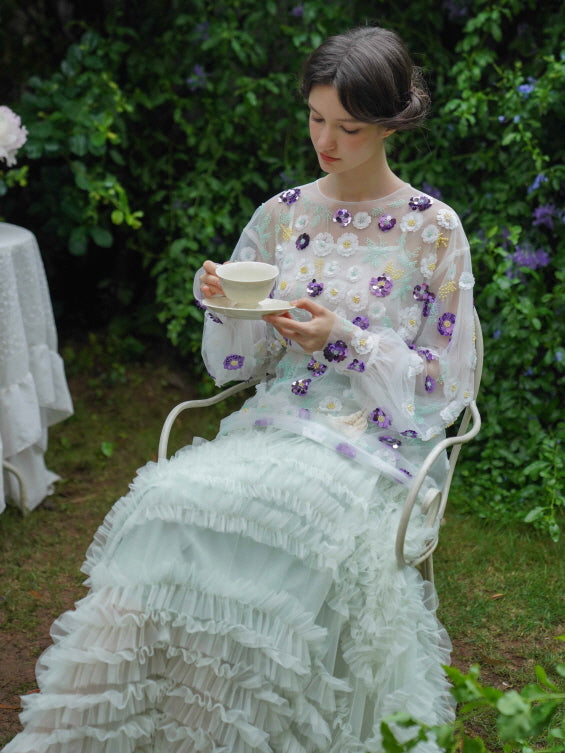 The model enjoys tea in a sheer floral embroidered top with a mint ruffle skirt, blending vintage charm and fairycore elegance in the garden.