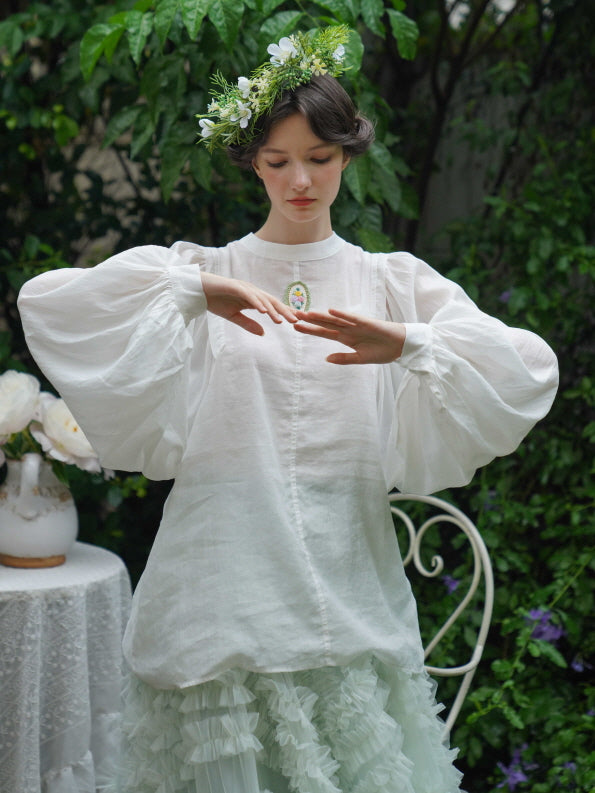 The model poses serenely in a white blouse with voluminous sleeves and a floral brooch, paired with a mint ruffle skirt for a fairycore garden look.
