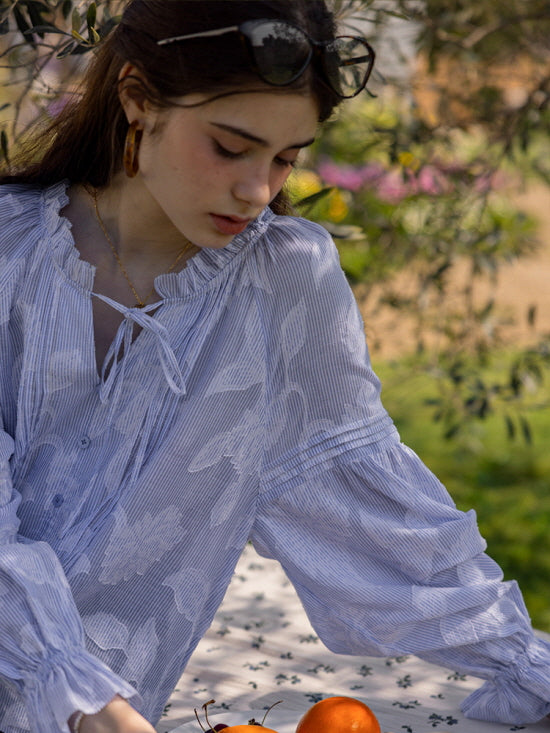 A young woman wearing a blue and white floral-patterned blouse with a ruffled neckline and tie detail, standing outdoors with a calm expression. The cottagecore-inspired look exudes a serene, fairytale vibe.