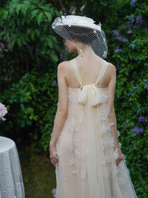 Back view of a romantic beige tulle dress with a long chiffon bow, as the model stands in a flower-filled garden wearing a vintage white hat.