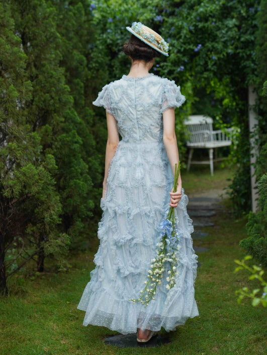 Back view of a woman walking through a fairytale garden in a frilled pastel dress, holding flowers with a soft vintage and cottagecore touch.