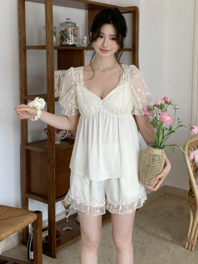 Model holding flowers while wearing a soft white coquette pajama set with lace trim and a layered top, styled in a warm cottagecore setting.